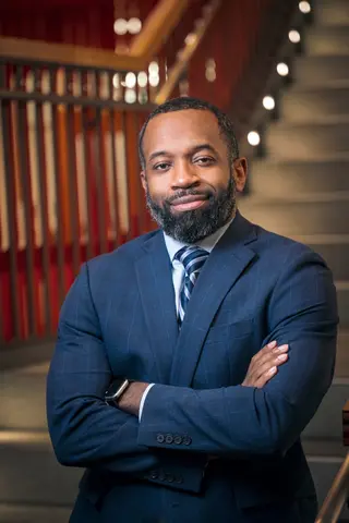 A man in a navy suit is smiling and looking at the camera, standing with his arms folded in front of stairs in the Student Experience Center
